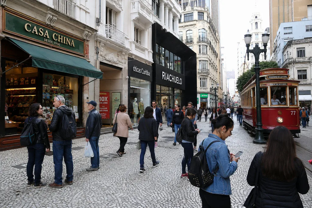 Fachada de uma antiga loja familiar no centro de Curitiba ao lado de uma grande rede varejista moderna, com consumidores utilizando smartphones para compras online.