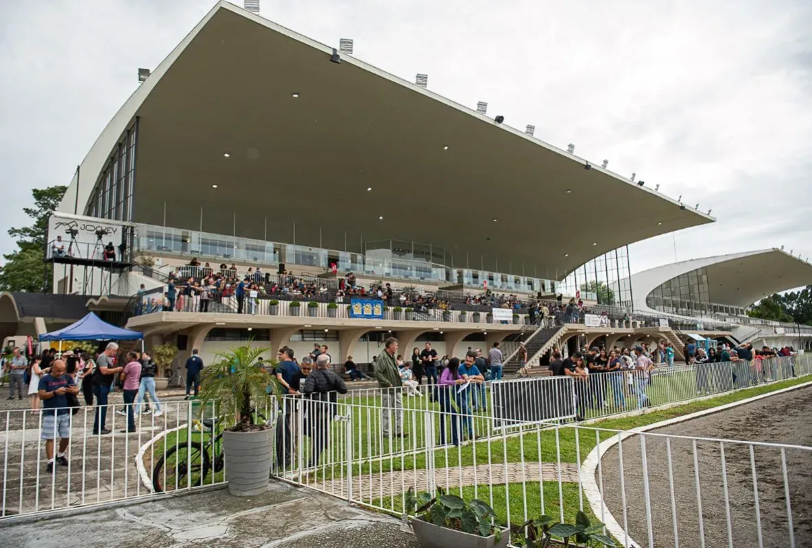 Pessoas assistindo corridas no Jockey Club do Paraná com arquibancada e área ao ar livre.