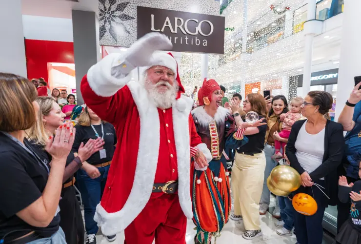 Papai Noel acenando em evento no shopping, cercado por pessoas e personagem vestido de palhaço.