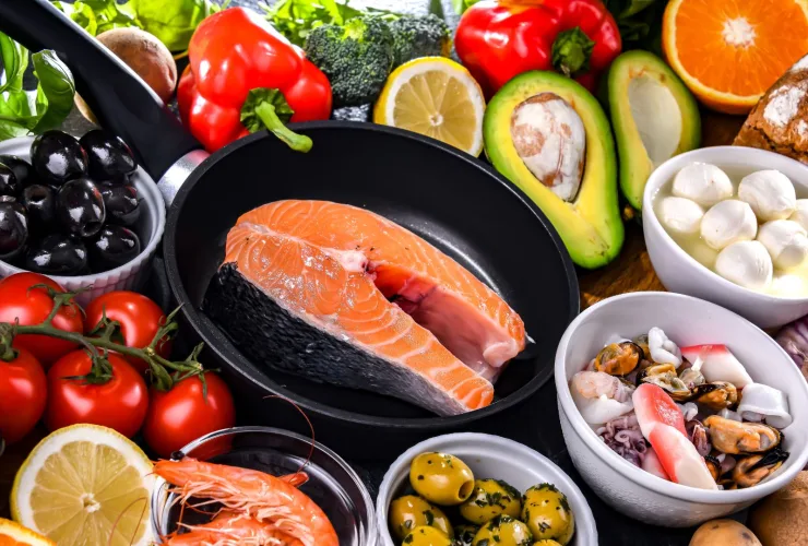 Posta de salmão em uma frigideira cercada por tomates, azeitonas, abacate, pimentões, limão, laranja, pão integral, queijo e frutos do mar