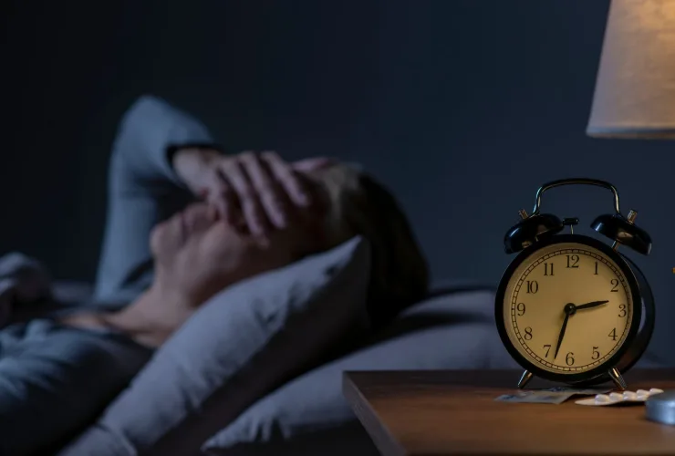 Pessoa deitada na cama, no escuro, com as mãos no rosto, ao lado de um despertador e de um abajur aceso