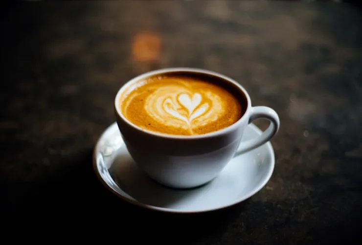 Xícara branca de café com desenho de coração na espuma, sobre um pires, em fundo escuro