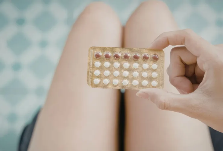 Mão segura uma cartela de pílulas anticoncepcionais, com as pernas de uma mulher sentada ao fundo