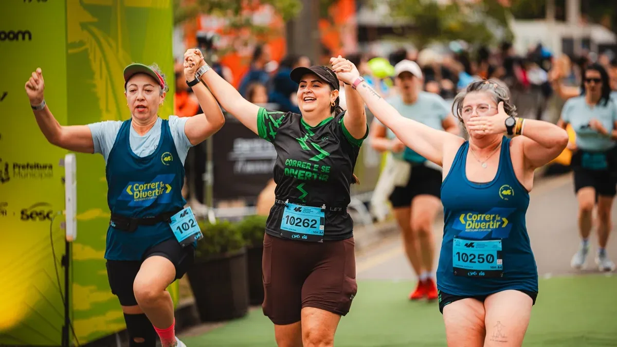 Três mulheres comemoram de mãos dadas durante corrida de rua, com outras pessoas ao fundo.