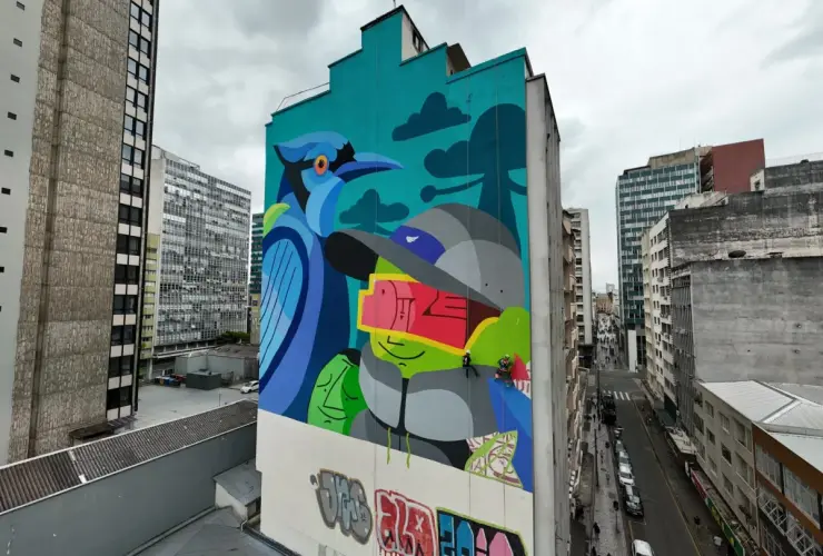 Mural colorido em prédio alto no centro urbano, com pintura de gralha-azul e figura humana estilizada, artistas trabalhando suspensos.