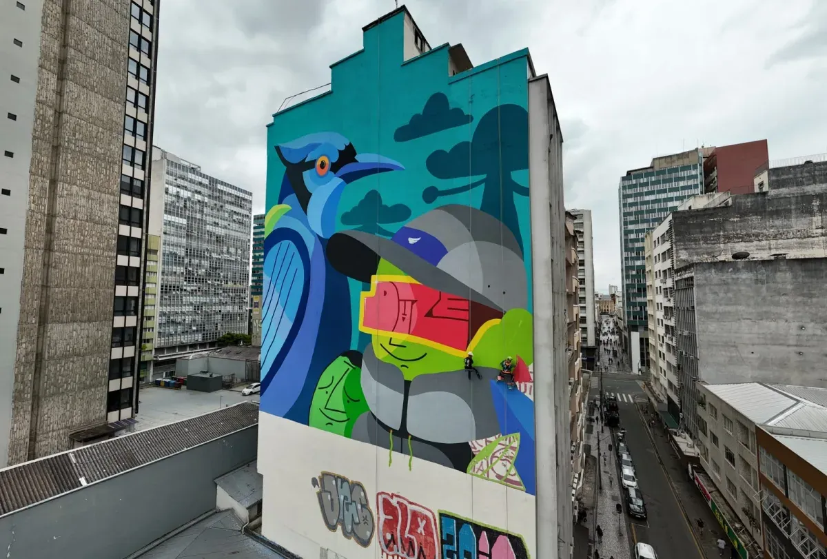 Mural colorido em prédio alto no centro urbano, com pintura de gralha-azul e figura humana estilizada, artistas trabalhando suspensos.