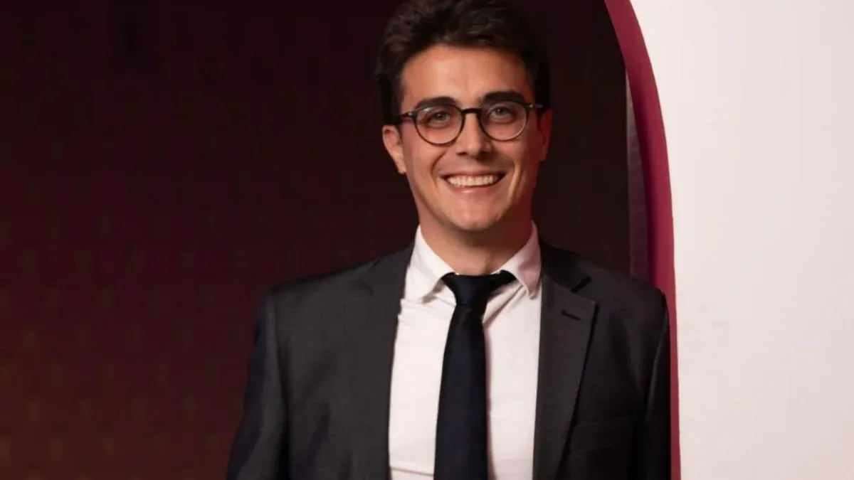 Homem sorridente de terno, camisa branca e gravata preta, usando óculos, posando em ambiente interno com fundo escuro e parede branca curva.