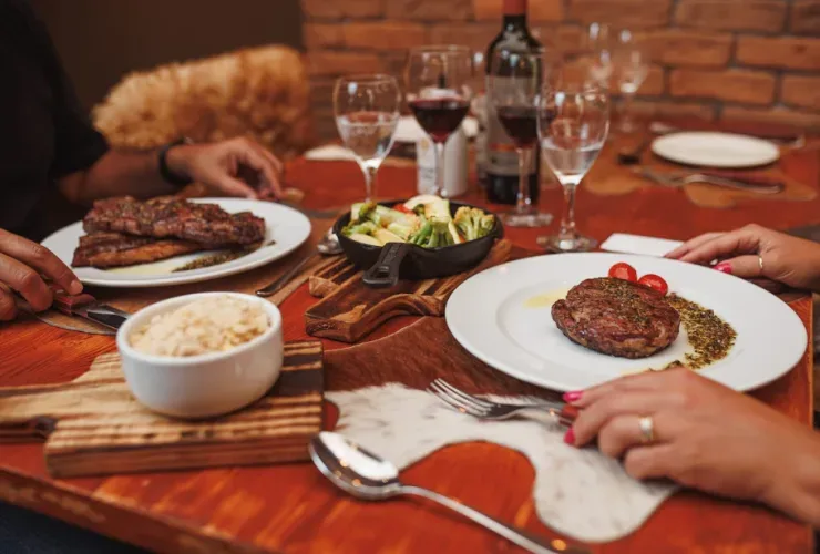 Mesa de madeira com pratos de carne, arroz, legumes, taças de vinho e garrafa de vinho tinto, com duas pessoas à mesa.