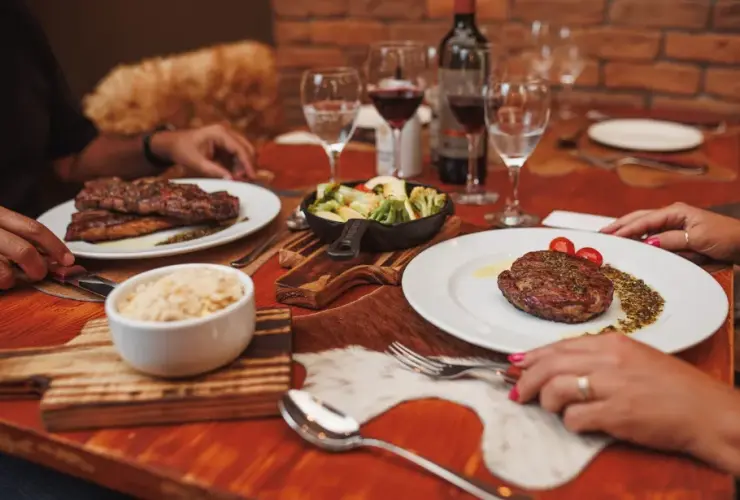 Mesa de madeira com pratos de carne, arroz, legumes, taças de vinho e garrafa de vinho tinto, com duas pessoas à mesa.