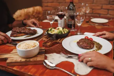 Mesa de madeira com pratos de carne, arroz, legumes, taças de vinho e garrafa de vinho tinto, com duas pessoas à mesa.