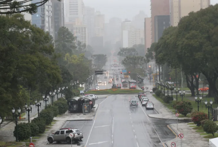 Curitiba sob chuva intensa, com nuvens carregadas, ruas molhadas e vegetação urbana.