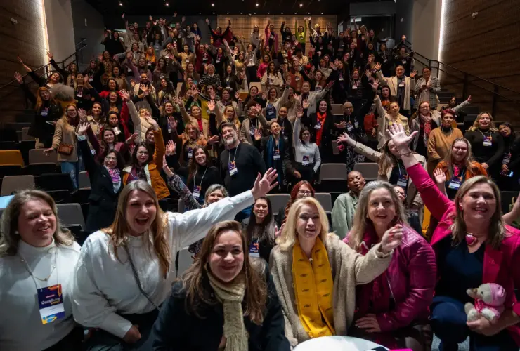 Grupo de pessoas em auditório sorrindo e acenando durante evento de empreendedorismo feminino.