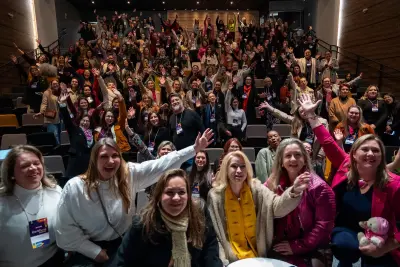 Grupo de pessoas em auditório sorrindo e acenando durante evento de empreendedorismo feminino.