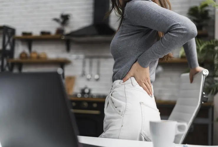 Mulher com a mão na lateral do quadril, apoiada em uma cadeira ao lado de uma mesa.