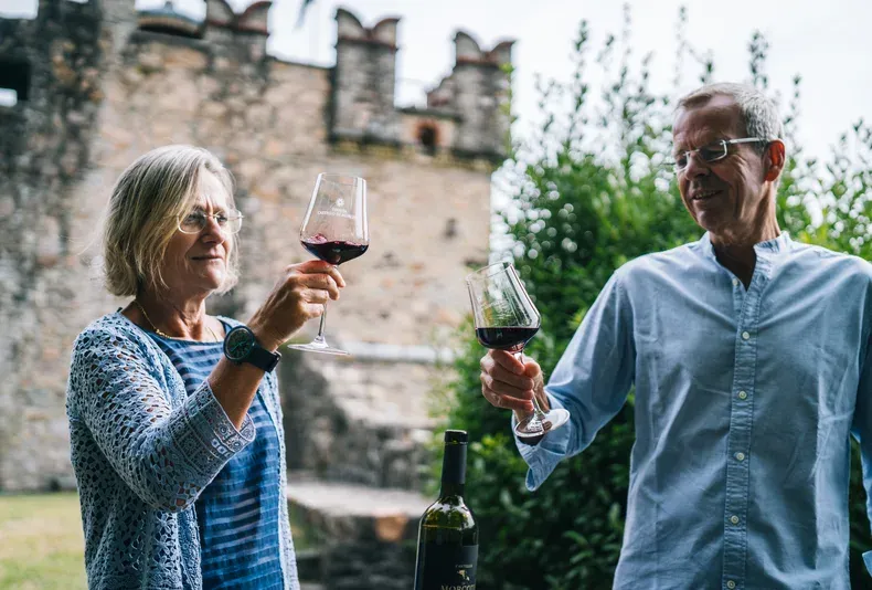 Casal adulto brinda com taças de vinho tinto ao ar livre, com muro de pedra ao fundo.