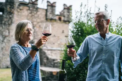 Casal adulto brinda com taças de vinho tinto ao ar livre, com muro de pedra ao fundo.
